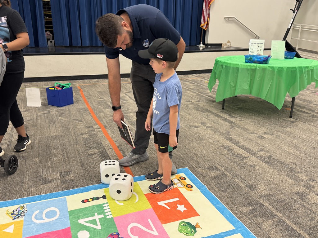 A man and a child play on a colorful mat with dice, standing in a room with blue curtains.
