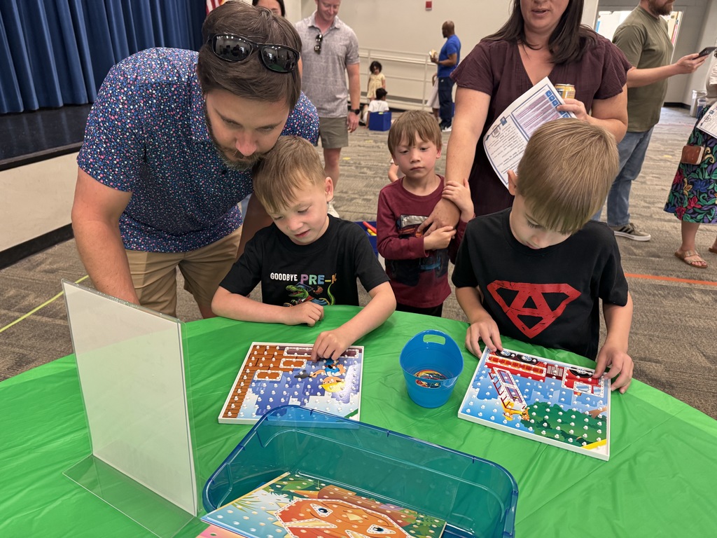 A man and two boys work on a board game at a table in a room with other families.
