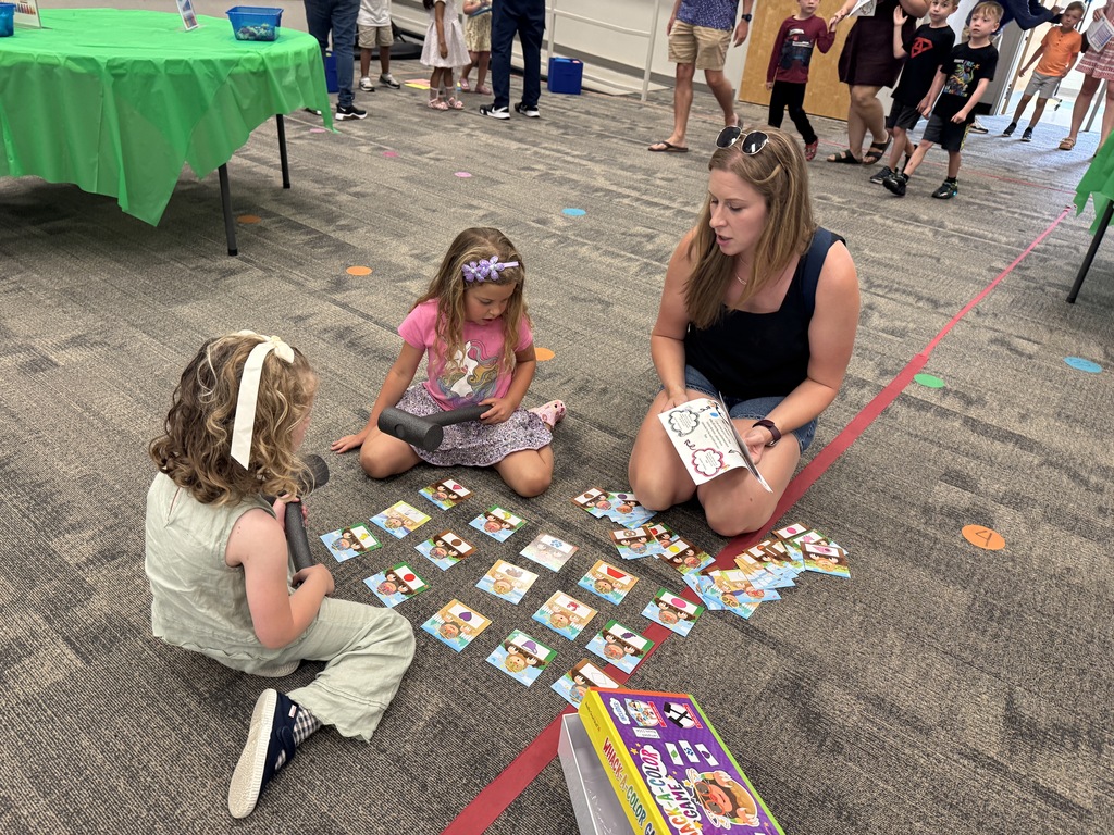 Two girls and a woman sit on the floor playing a matching game with cards. A table with a green tablecloth is in the background.
