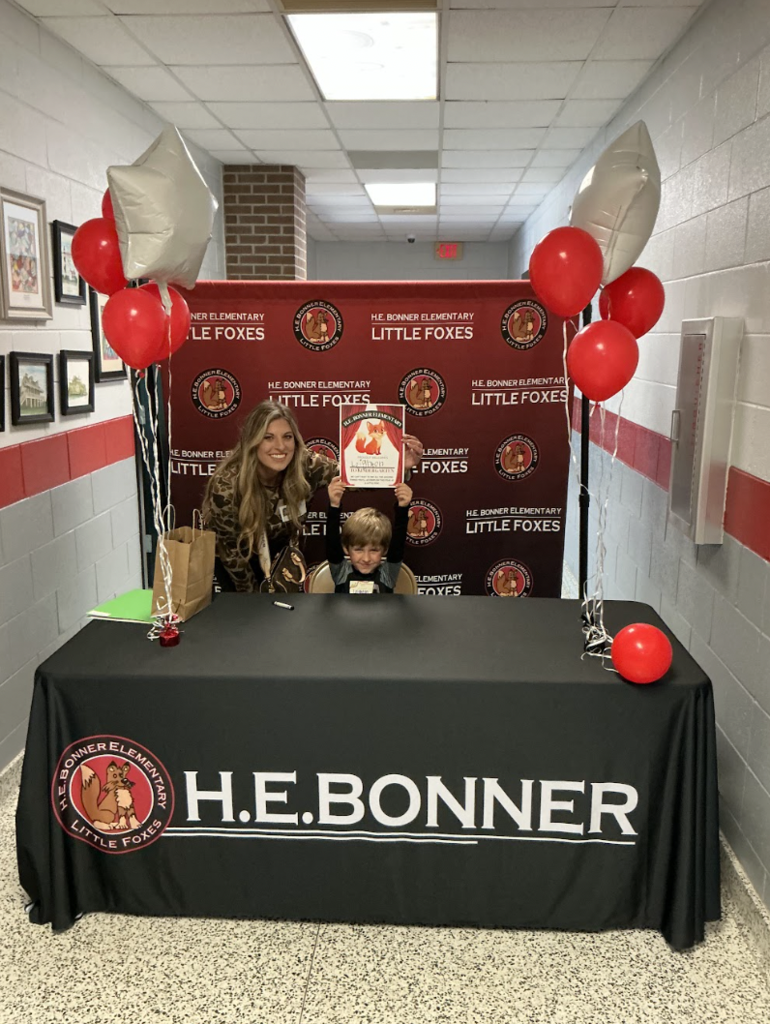 A child sitting at a table with a black table cloth that reads HE Bonner and the student is holding up a piece of paper.