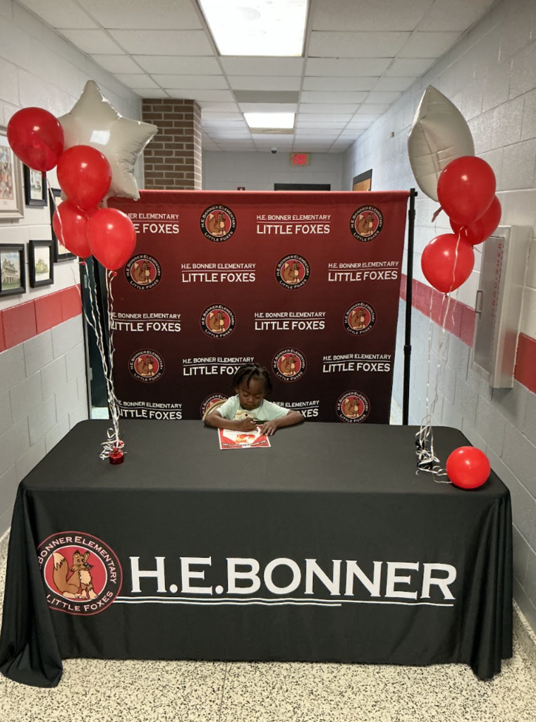 A person is seated at a table with a black tablecloth that reads "H.E. Bonner". Behind them, balloons and a red banner are displayed.