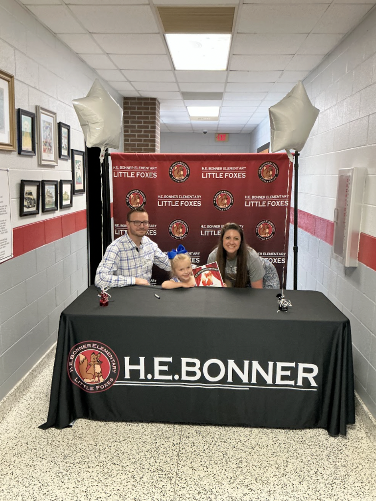 Family sits behind a desk with a sign reading "H.E. Bonner" in a hallway. A banner reads "Little Foxes" behind them.