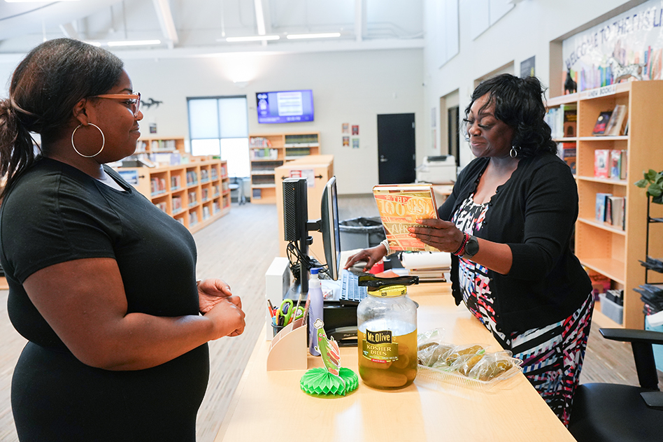 Two women stand in a library. One holds a book and stands behind a desk with jars.