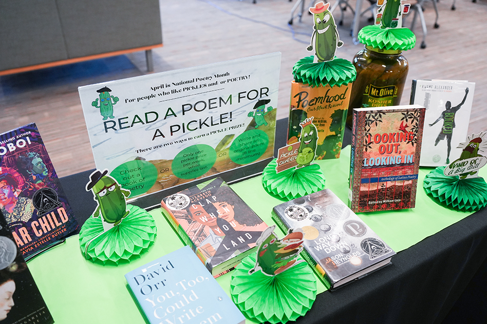Books on a table with green decorations. Sign reads "Read a Poem for a Pickle!" Titles include "Looking Out", "Robot", and "David Ortiz Comes to Town".