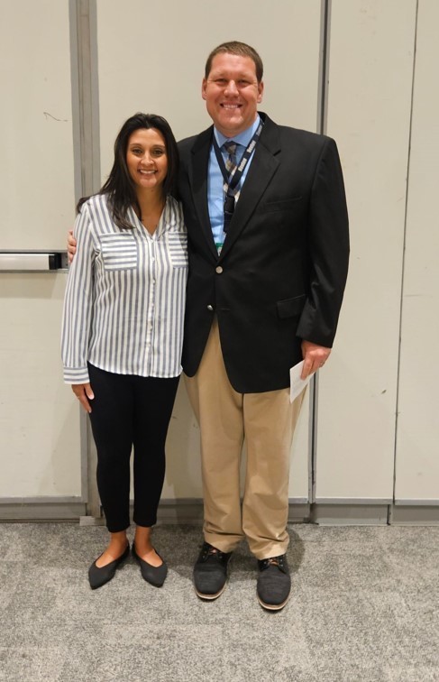 A man and woman in formal attire stand close, smiling, against a white wall. The man wears a tie.