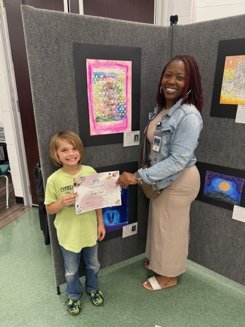 A young child holds a certificate while standing next to a woman who smiles in front of a display of artwork.