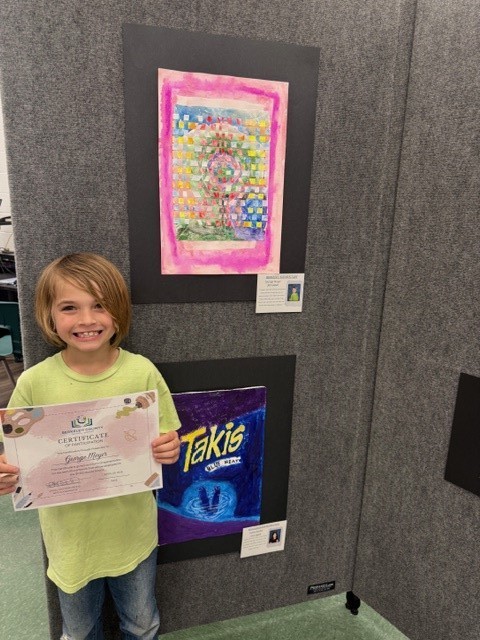 A child in a yellow shirt holds a certificate in front of a wall with two framed artworks.