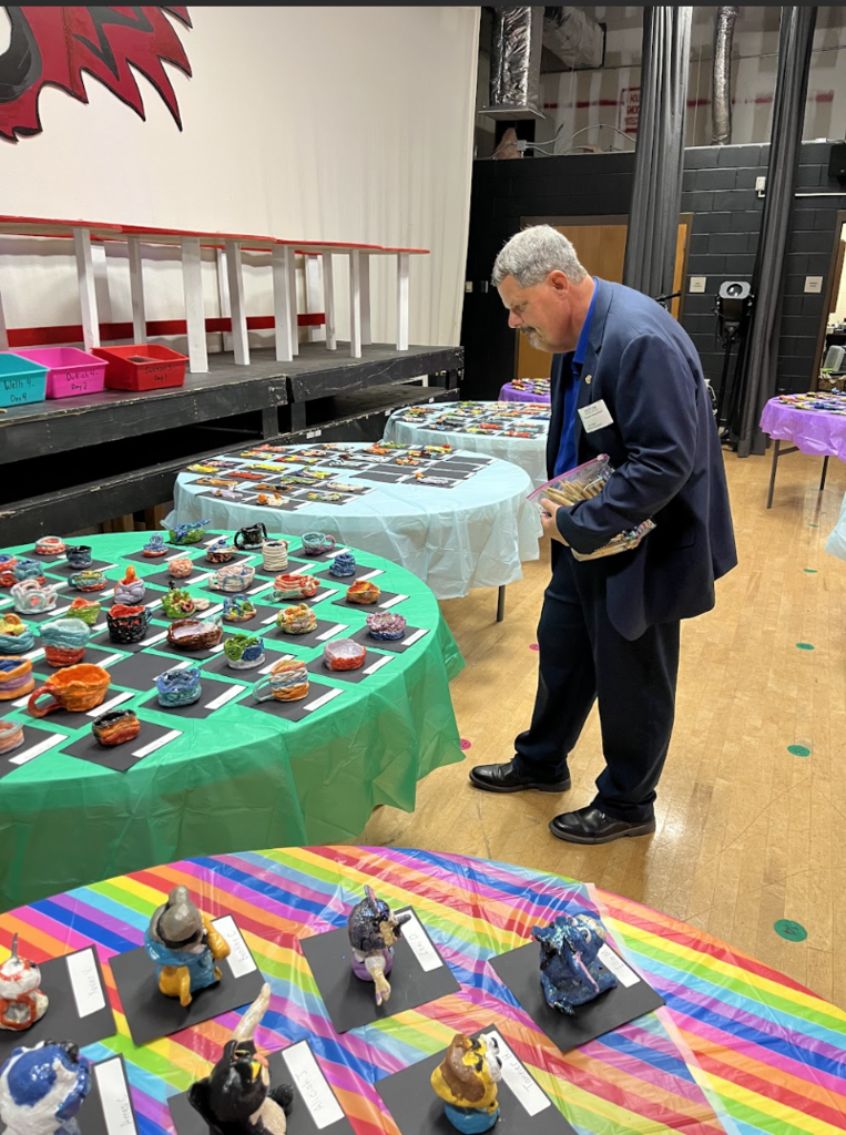 A man in a suit inspects colorful ceramic creations on a table with a green cloth.