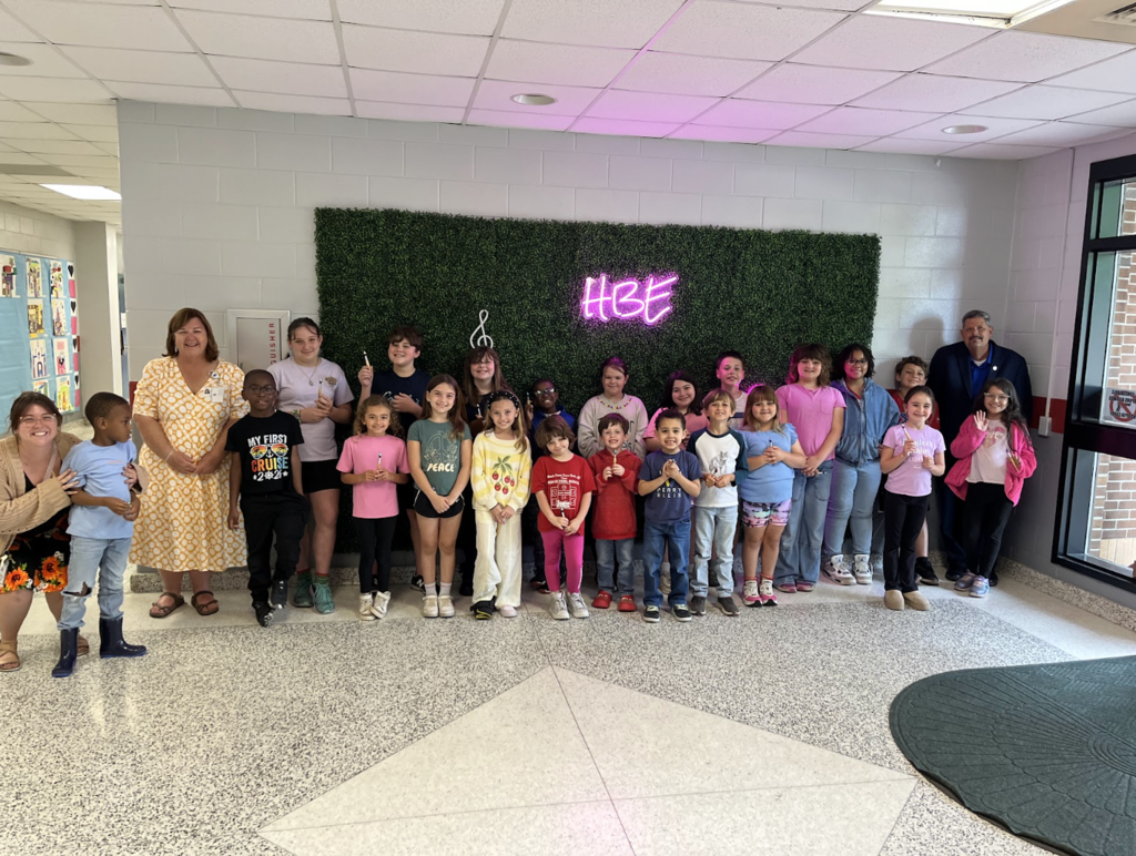 A group of young children and adults stand in a room. The wall behind them features a wall of greenery and a pink neon sign.
