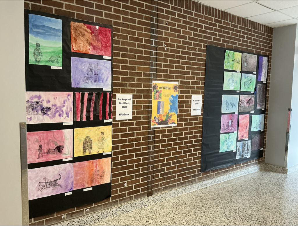 Multiple student artwork on display. A brick wall with white tiles. Light from a hallway visible at the bottom.