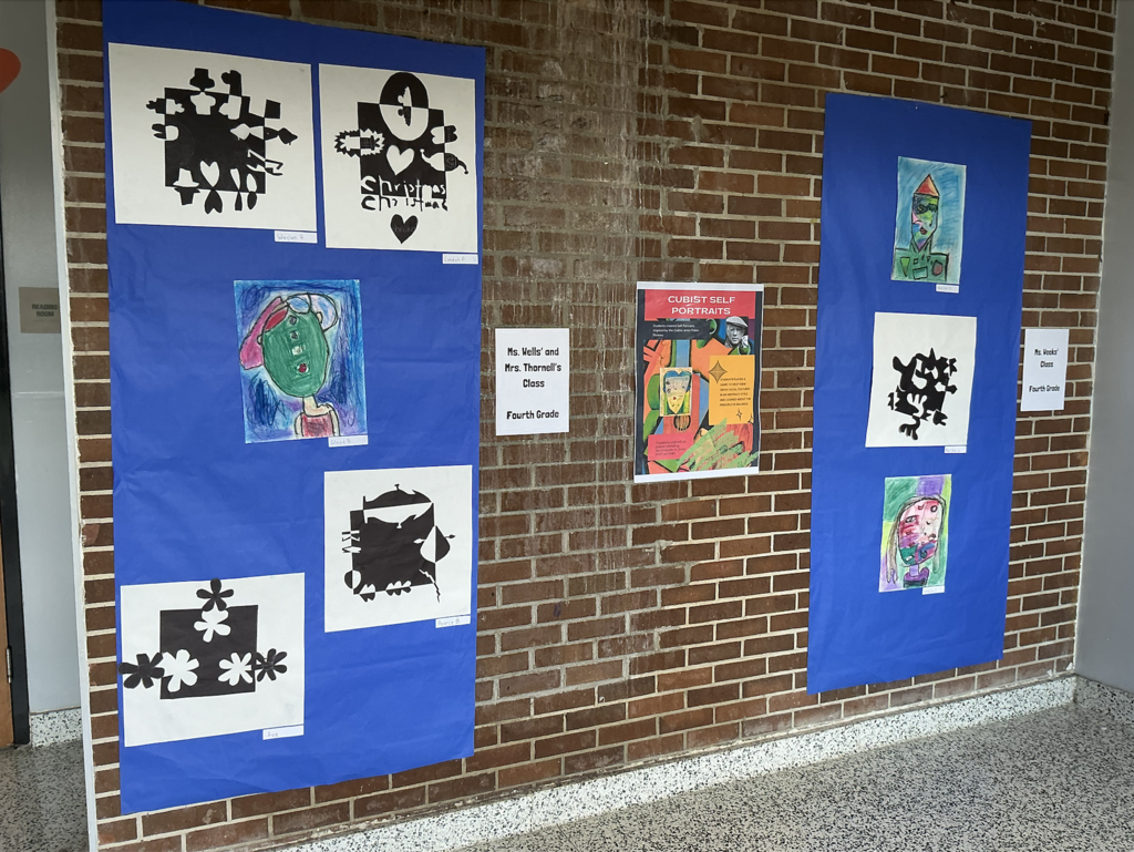 Blue backdrop with multiple artworks. Various shapes, colors, and styles are present. A brick wall with a gray floor.