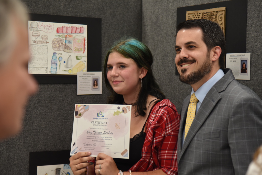 A young woman with green hair and a red shirt holds a certificate in front of a man in a suit.