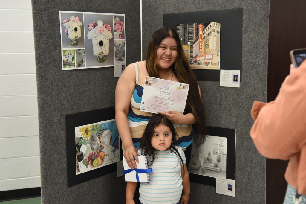 Woman and child standing behind a display of art. Woman holding a certificate. Child holding a wrapped gift.