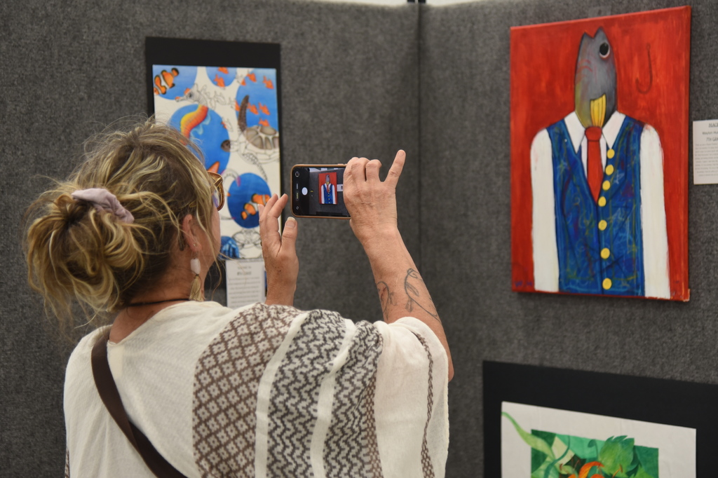 A person takes a photo of a painting depicting a man in a suit. Nearby, other paintings are on display.