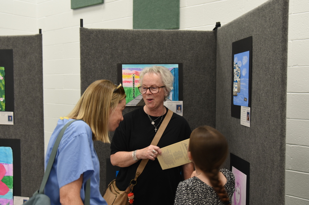 Three women are in a room with artwork on the walls, one holding paper and speaking.