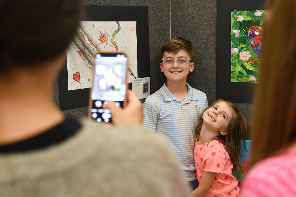 A person takes a photo with a cell phone of two smiling children in front of art displays.