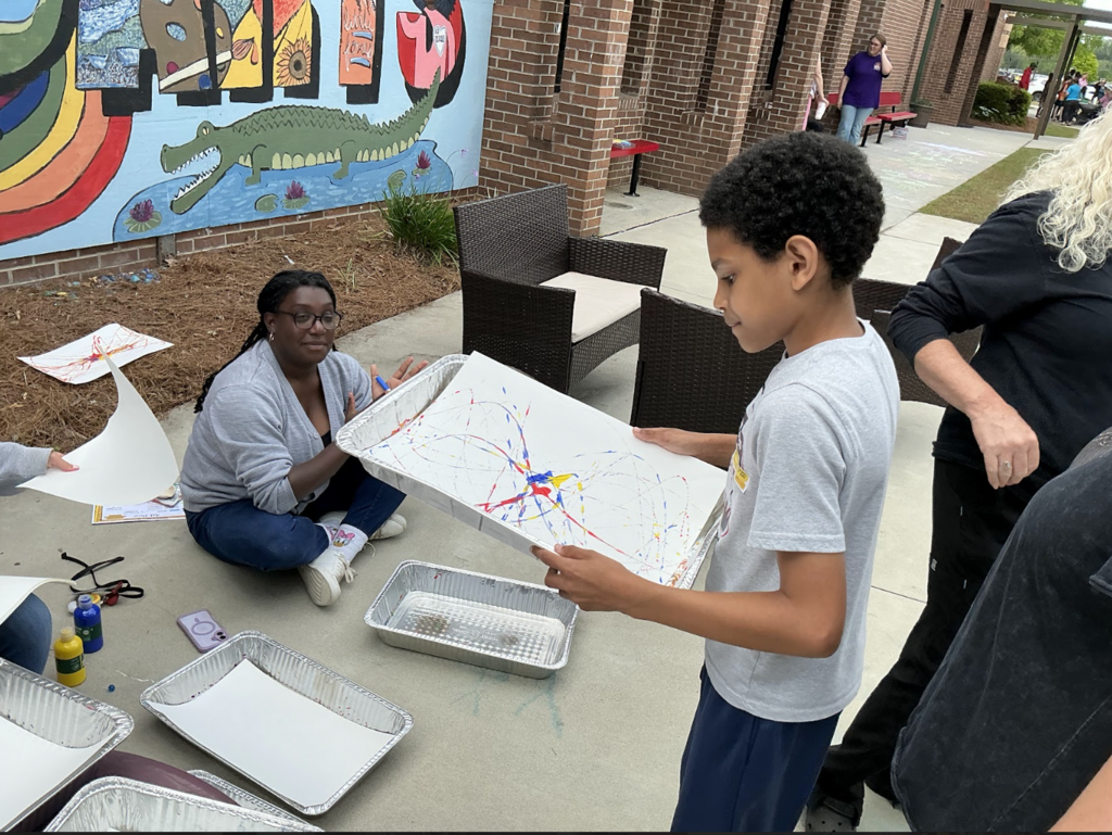 A young boy holds a colorful painting while a woman sits nearby on the ground. A mural of a rainbow and alligator is visible behind them.