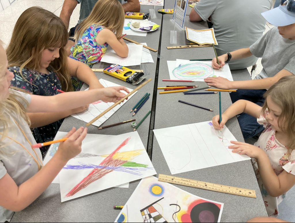 Children seated around a table, each engaged in drawing with crayons on paper. Various colored drawings and art supplies are on the table.