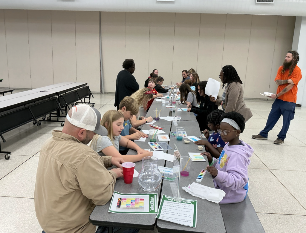Several children in a classroom setting, seated at long tables with papers and art supplies. An adult sits at the end.