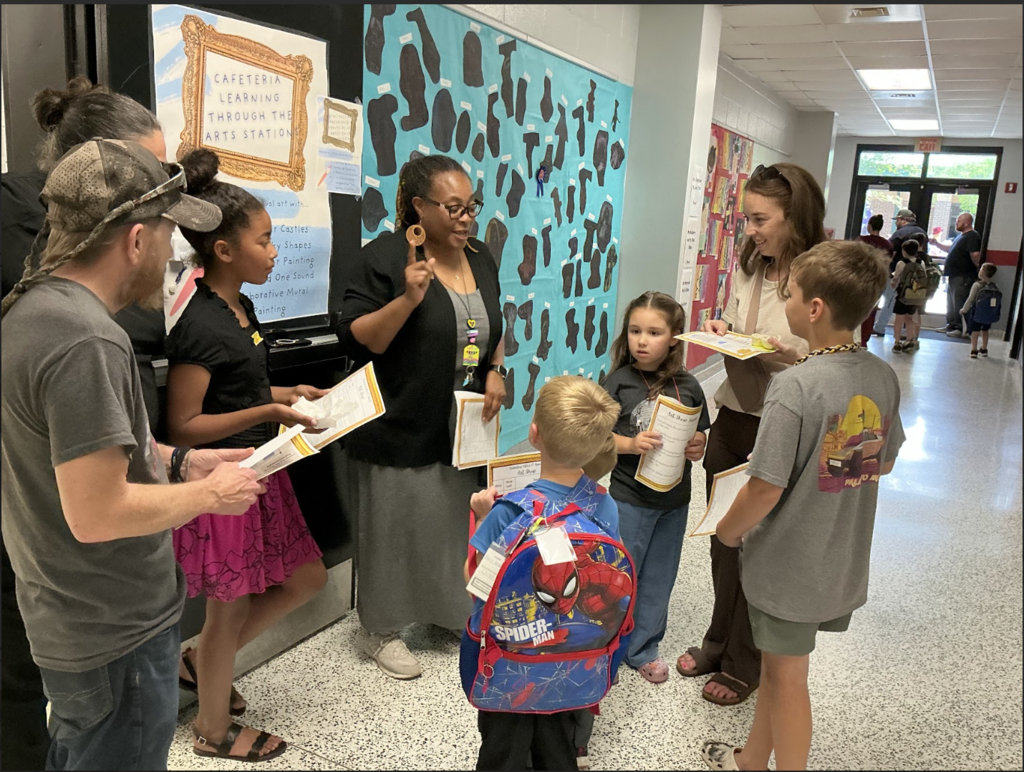People stand in a hallway. A man reads from a paper to a group of children.