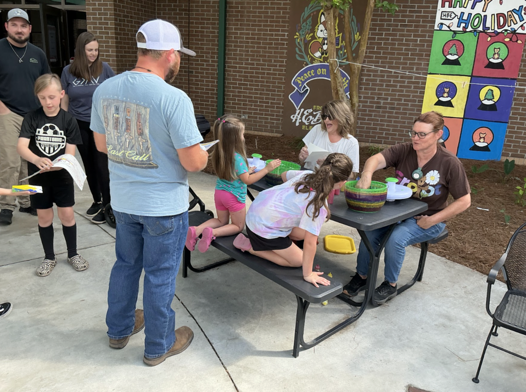 People gather at a table outside, with some seated and others standing. A banner with colorful squares is in the background.