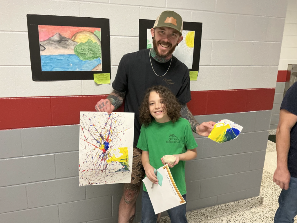 A man and a child pose with colorful artwork in a school hallway, three framed paintings on the wall.