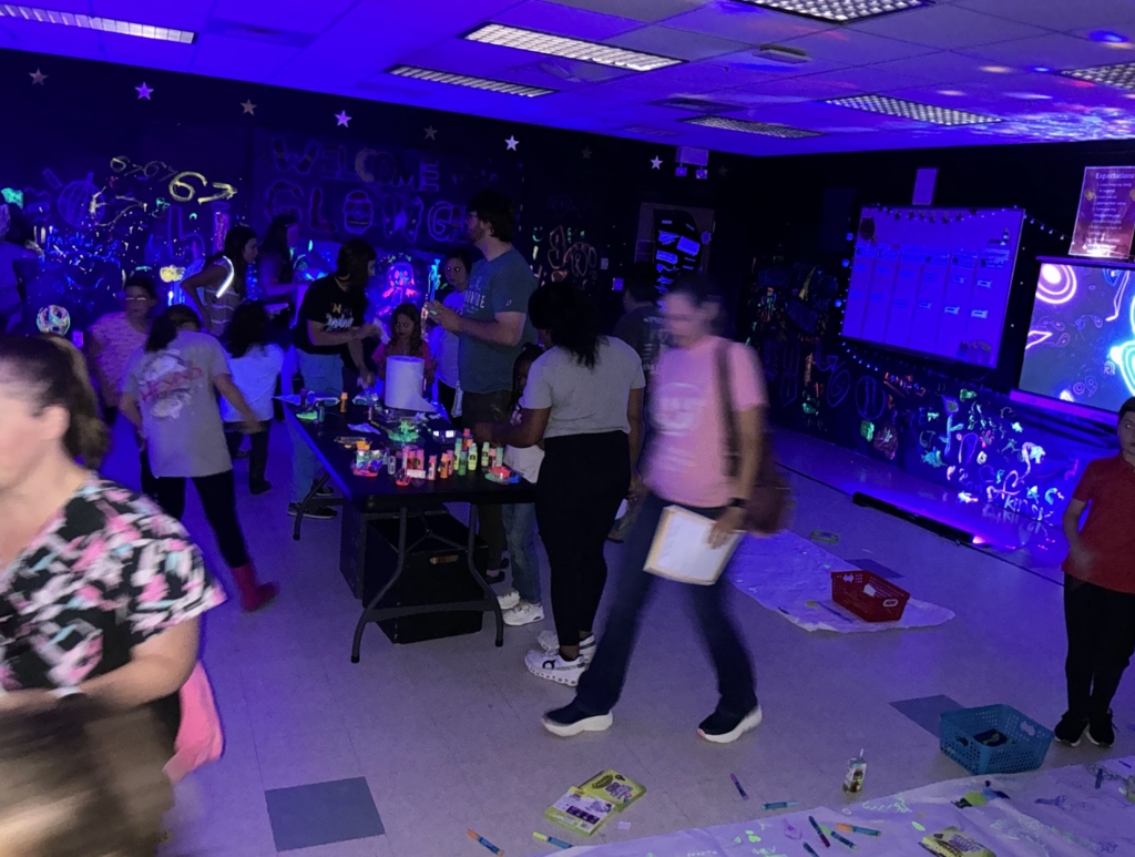 A room with a purple light setup, walls decorated with glowing art. People are walking around and one holds a folder.