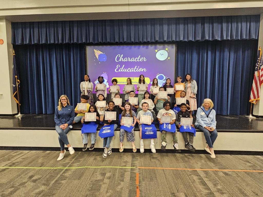 Children sit on a stage holding certificates, with two women, one seated and one standing. Behind them, a blue curtain and projector screen.