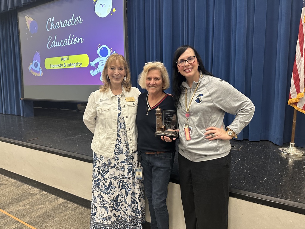 Three women stand on a stage, one holding a trophy, with a projector screen behind them displaying "Character Education."