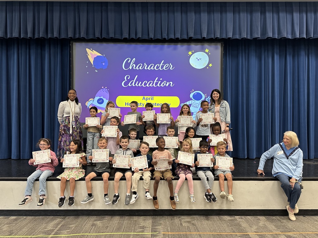 Students sit on stage holding certificates; two adults stand behind them. The stage is under a blue curtain with a projector screen in the background.