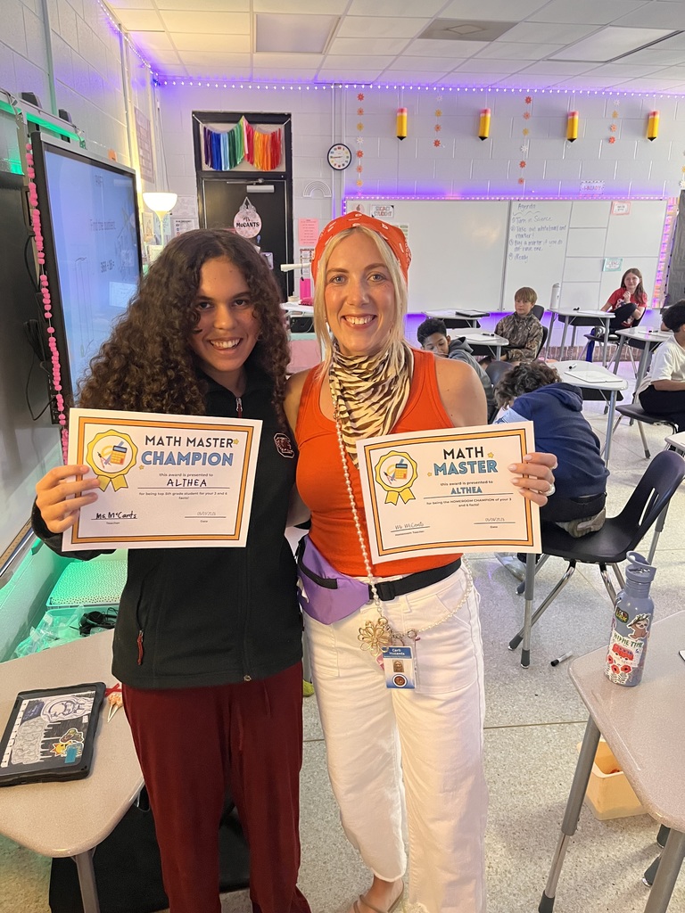 Two women in a classroom with colorful decorations hold certificates, one labeled Math Master Champion.