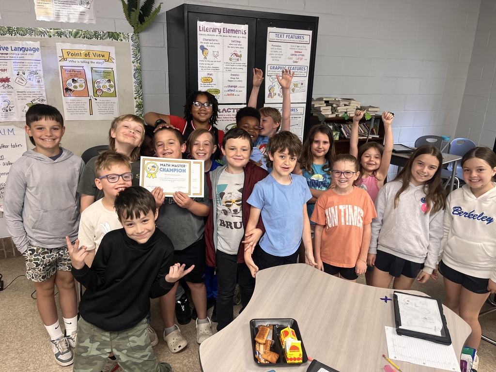 Students stand in a classroom, posing for a photo. Some hold certificates. A table with food is in the foreground.