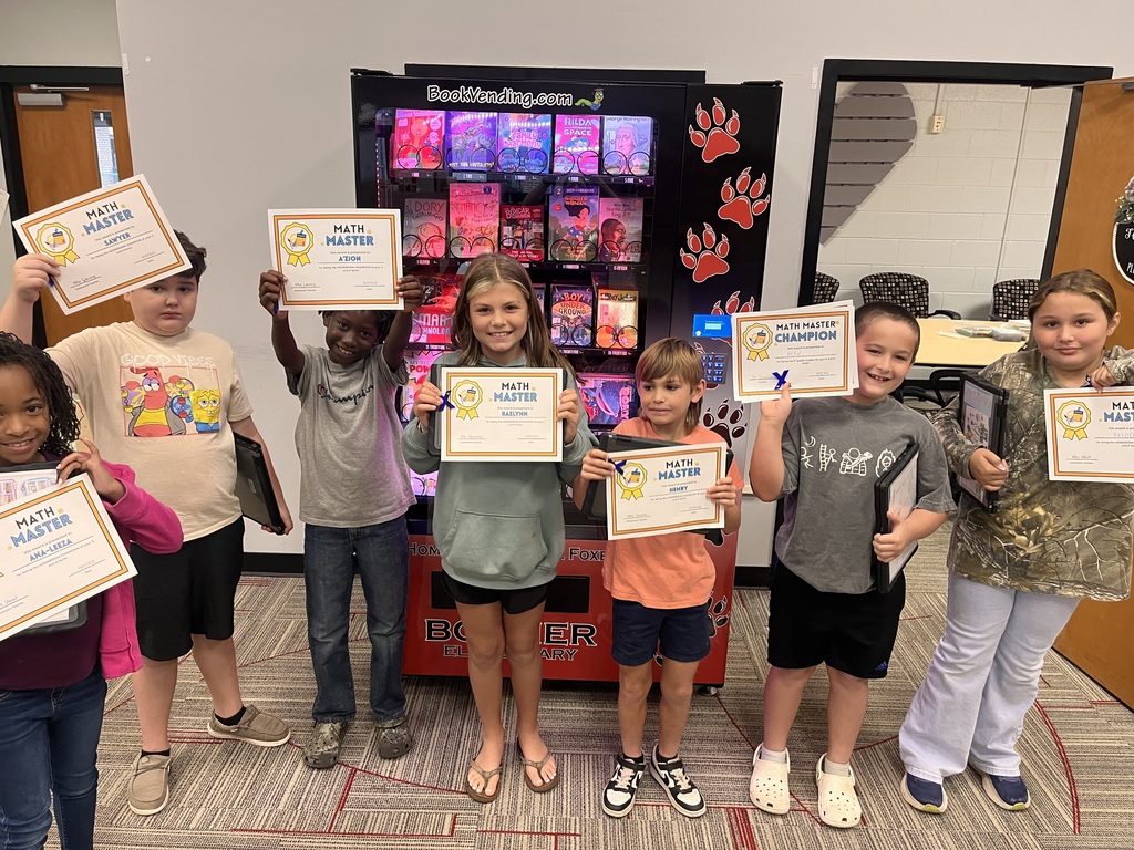 A group of children holding certificates stand in a room, with a vending machine displaying books in the background.