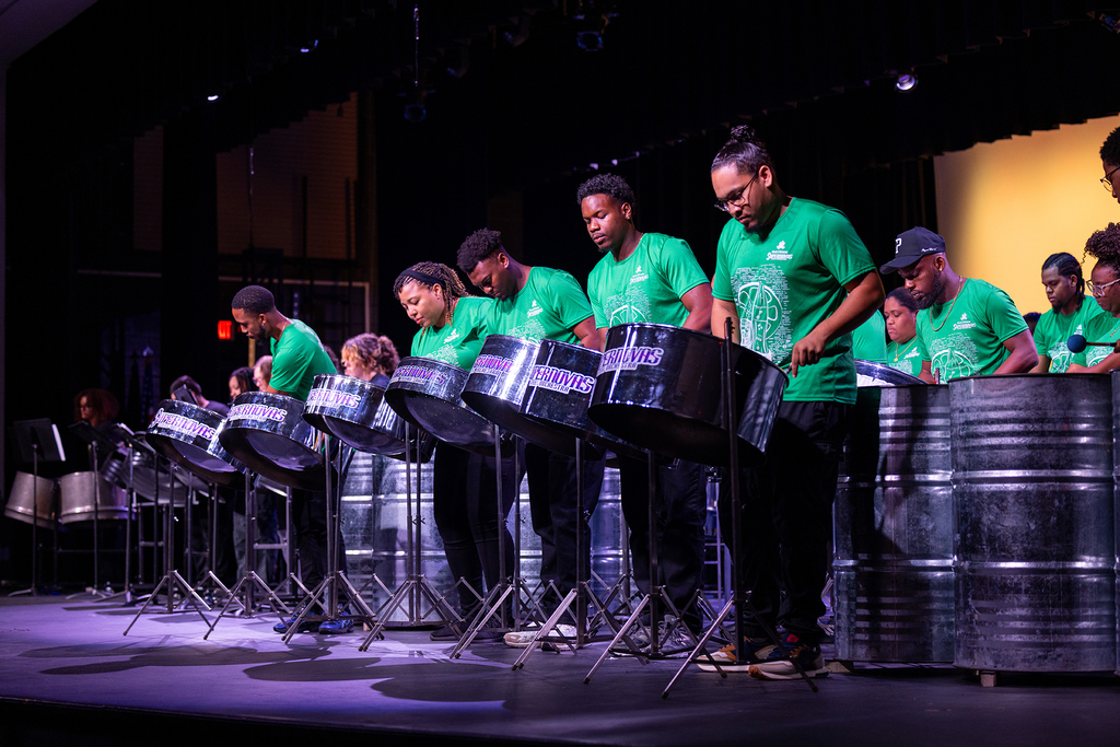 A group of people in green shirts play steel drums on a stage with a yellow backdrop.