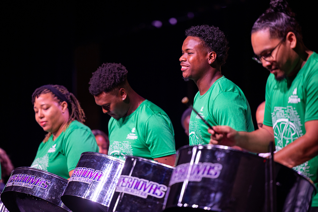 Four adults in green shirts stand behind drums with two adults playing steel drums. Background shows additional adults in green shirts.