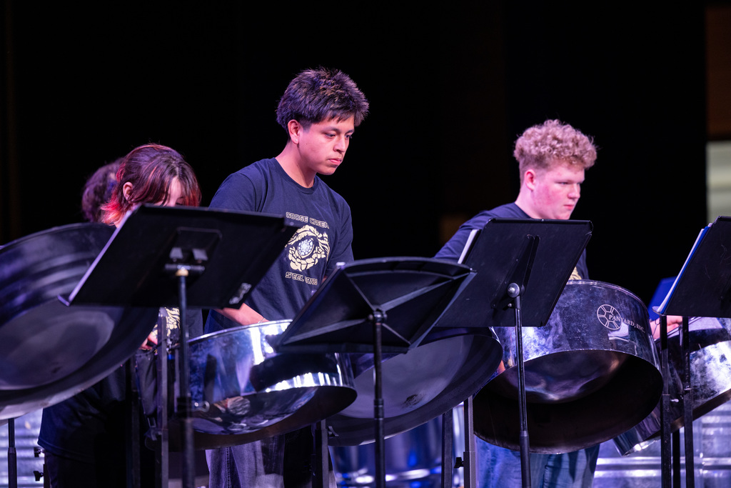 Four individuals, two boys and two girls, stand behind steel drums. They are likely performing at a musical event.