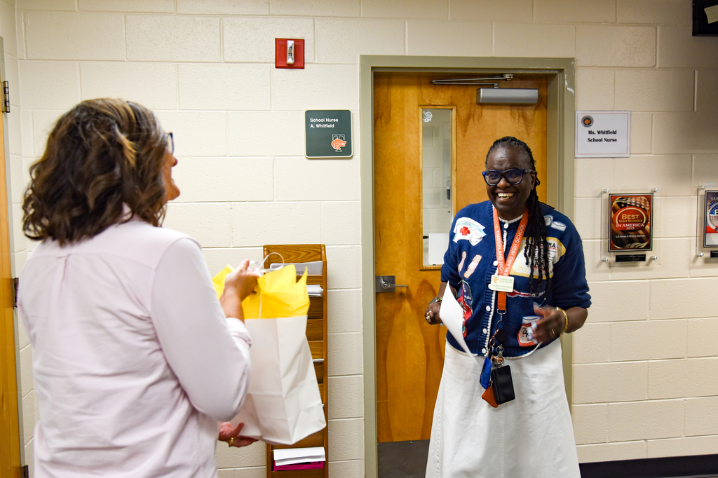 Two women stand in a hallway, one holding a bag, both engaged in conversation. Behind them is a white-painted room wall.