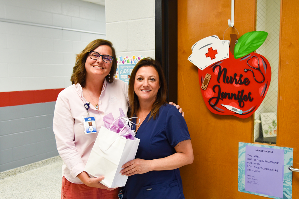 Two women stand in front of a door, one holding a gift bag. A red apple sign reads Nurse Jennifer.