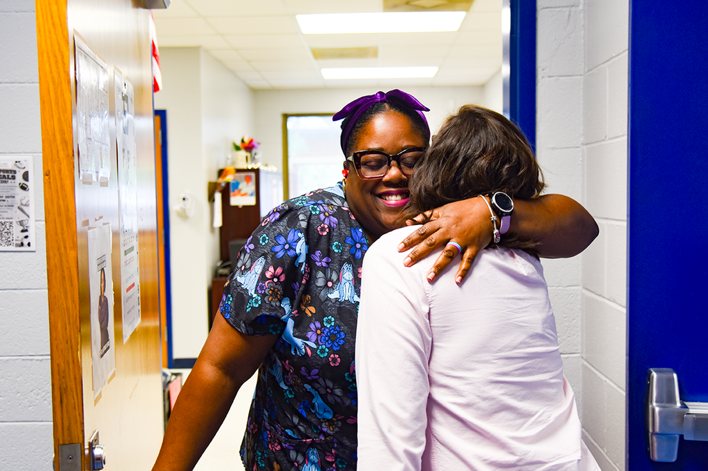 Two women stand in a hallway; one hugs the other, smiling and wearing glasses. Behind them, walls and doors are visible.