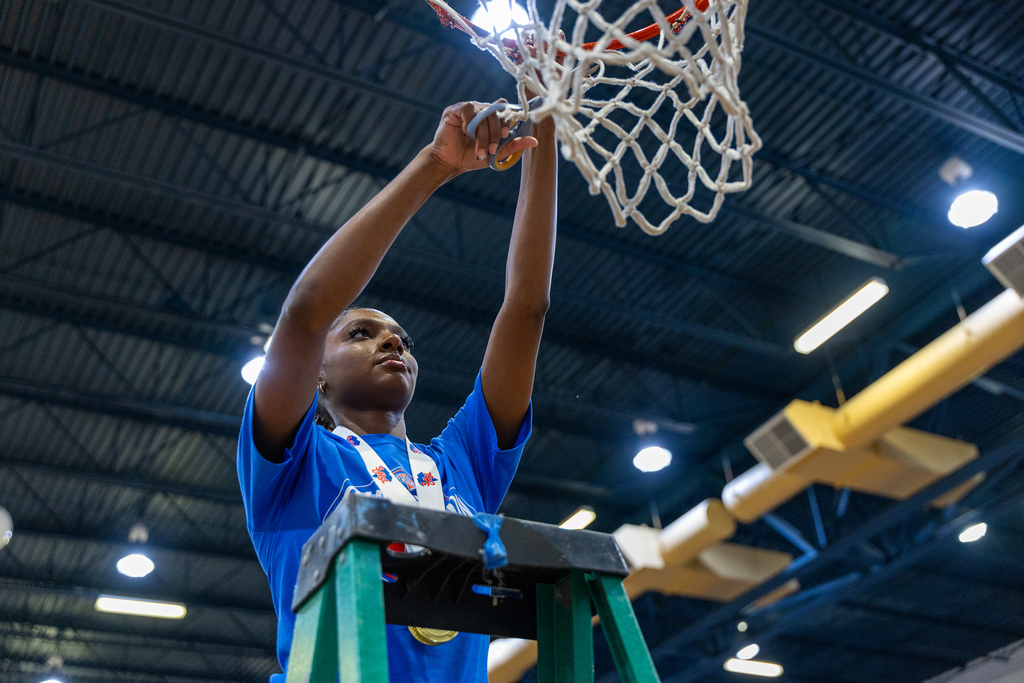 A person in a blue shirt on a ladder inside a gym, holding up a basketball net.