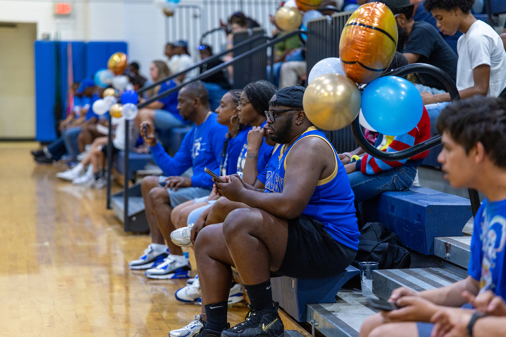 Spectators wearing blue shirts sit on bleachers of a basketball court, some holding cellphones.