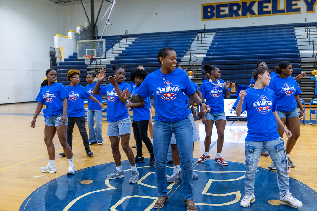 Nine women, dressed in blue tops and shorts, dance in a basketball court with Berkeley in the background.