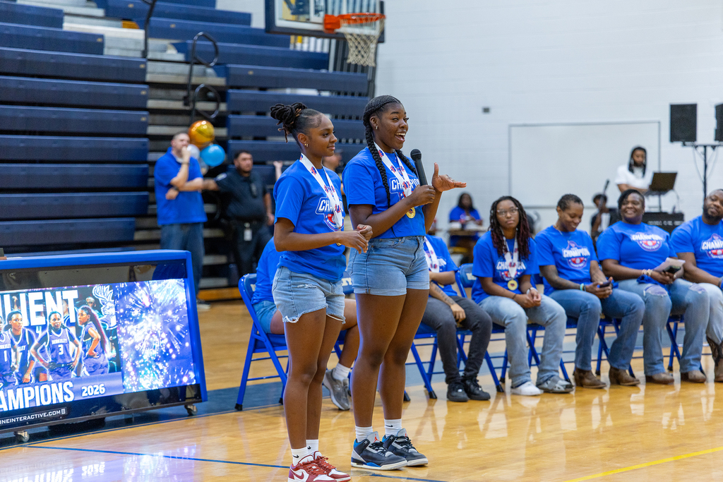 Two girls with medals stand on a basketball court while others in blue shirts sit and watch.