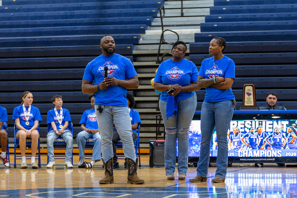 A group of people wearing blue shirts and jeans, standing in a gymnasium. A man speaks into a microphone.