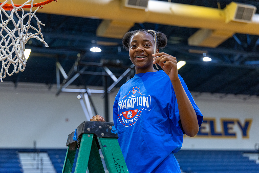 A woman wearing a blue shirt with "Champion" written on it stands on a ladder in a gym, smiling.