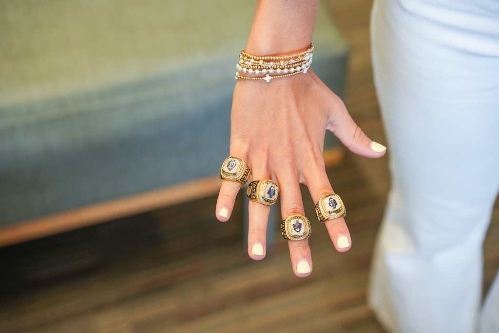 A close-up of a hand with three gold rings and a bracelet with pearls. The nails are painted yellow.
