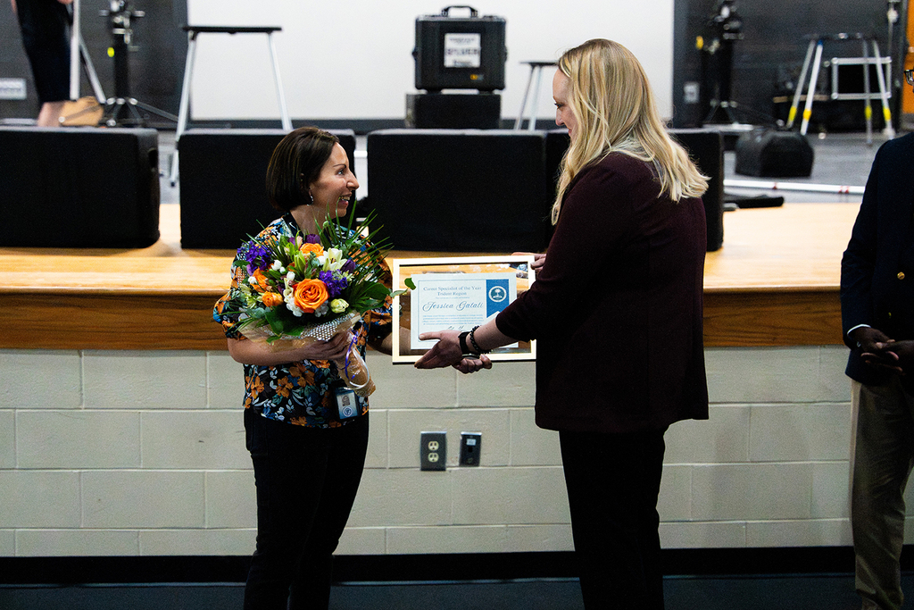 Two women stand on a stage. The woman in floral clothing holds a bouquet of flowers. The woman in a maroon coat holds a certificate.