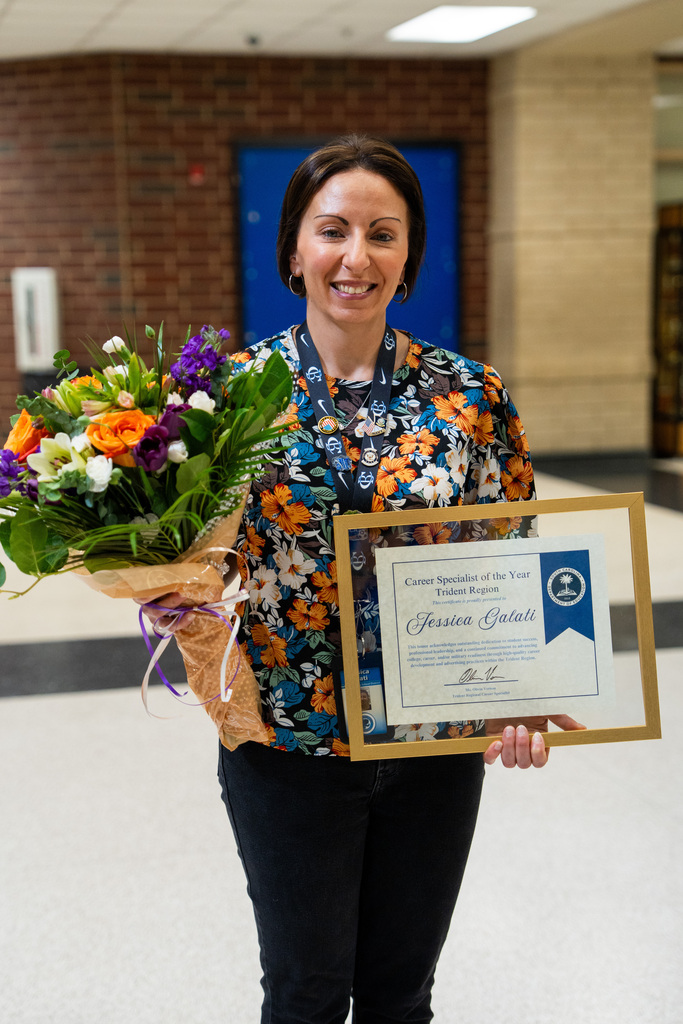 A woman in a colorful shirt stands with a bouquet of flowers and holds a framed certificate.