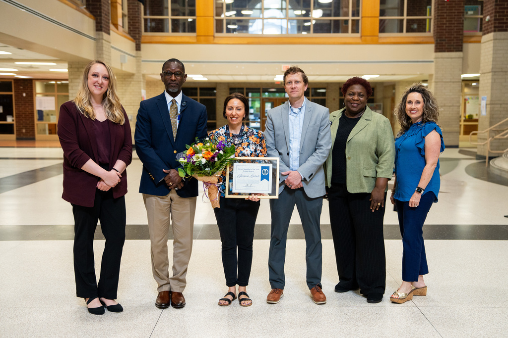 A group of people stands in a hallway. They hold a framed certificate. Two women wear sandals.