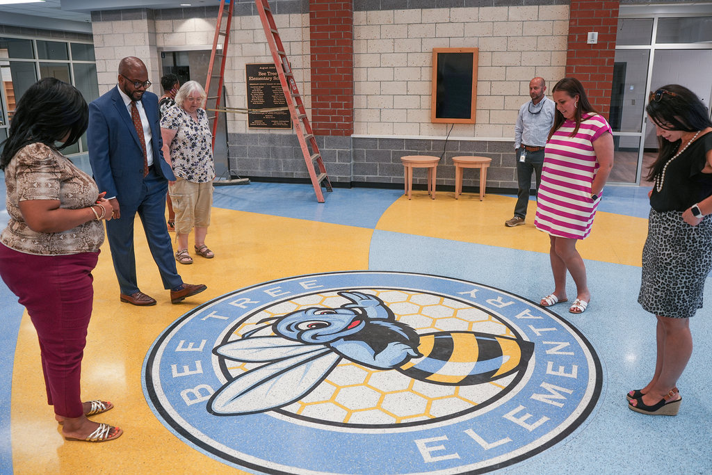 A group of adults stands around the Bee Tree Elementary logo on the floor of BTE.  Background includes brick walls and glass doors.